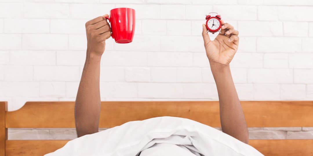 A person holding an alarm clock and a cup of coffee in bed