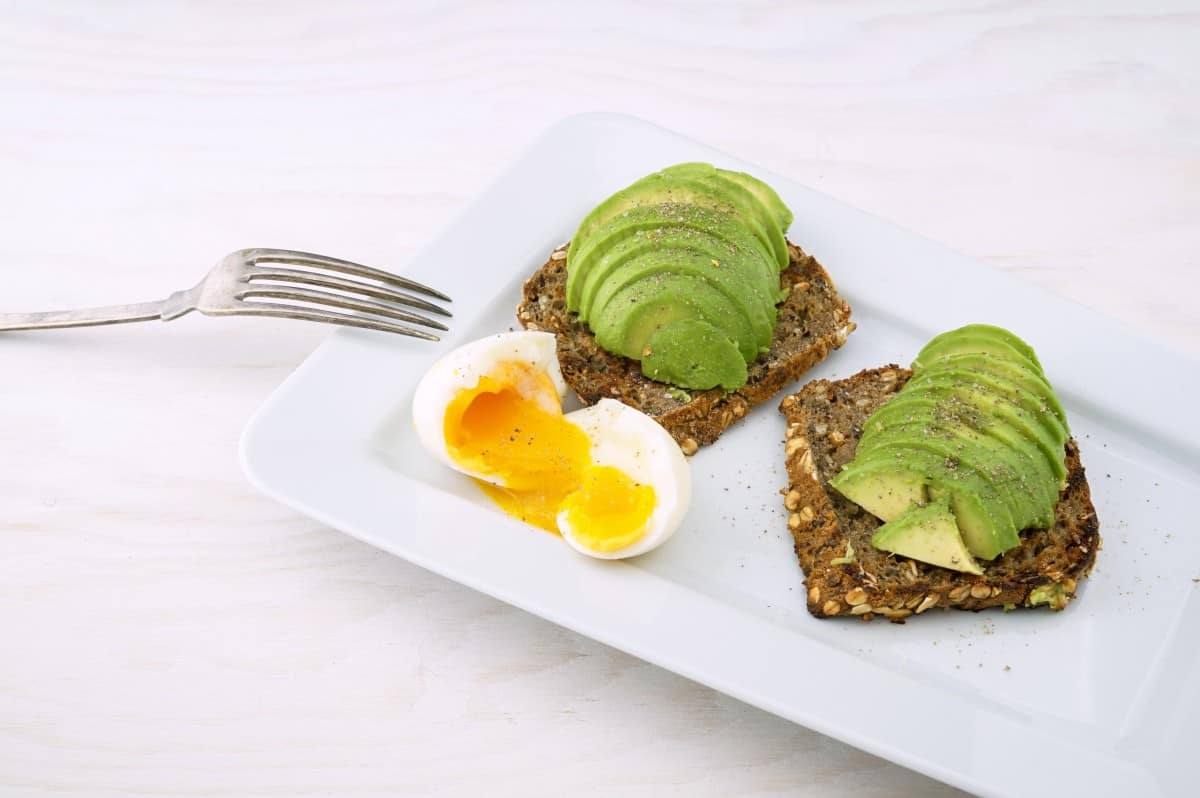 wholewheat bread avocado toast with soft boil egg on a white platter and fork on the side. 