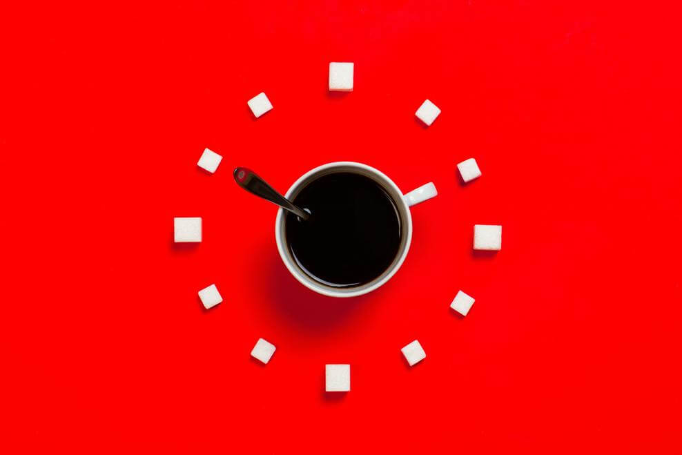A cup of black coffee surrounded by sugar cubes atop a red background.